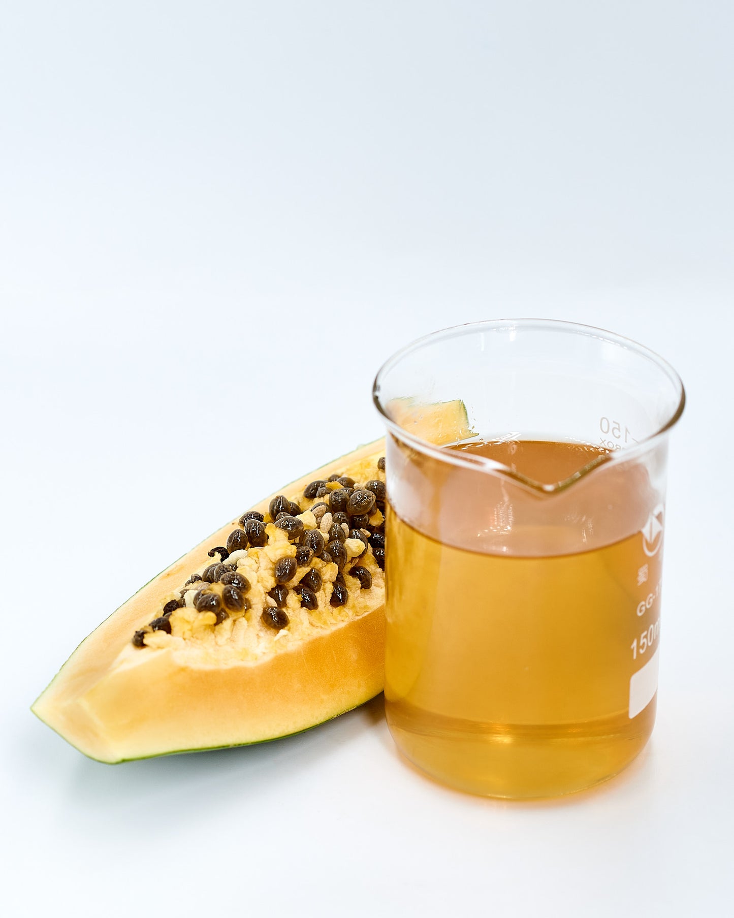 Glass of papaya oil next to a slice of papaya on a light background